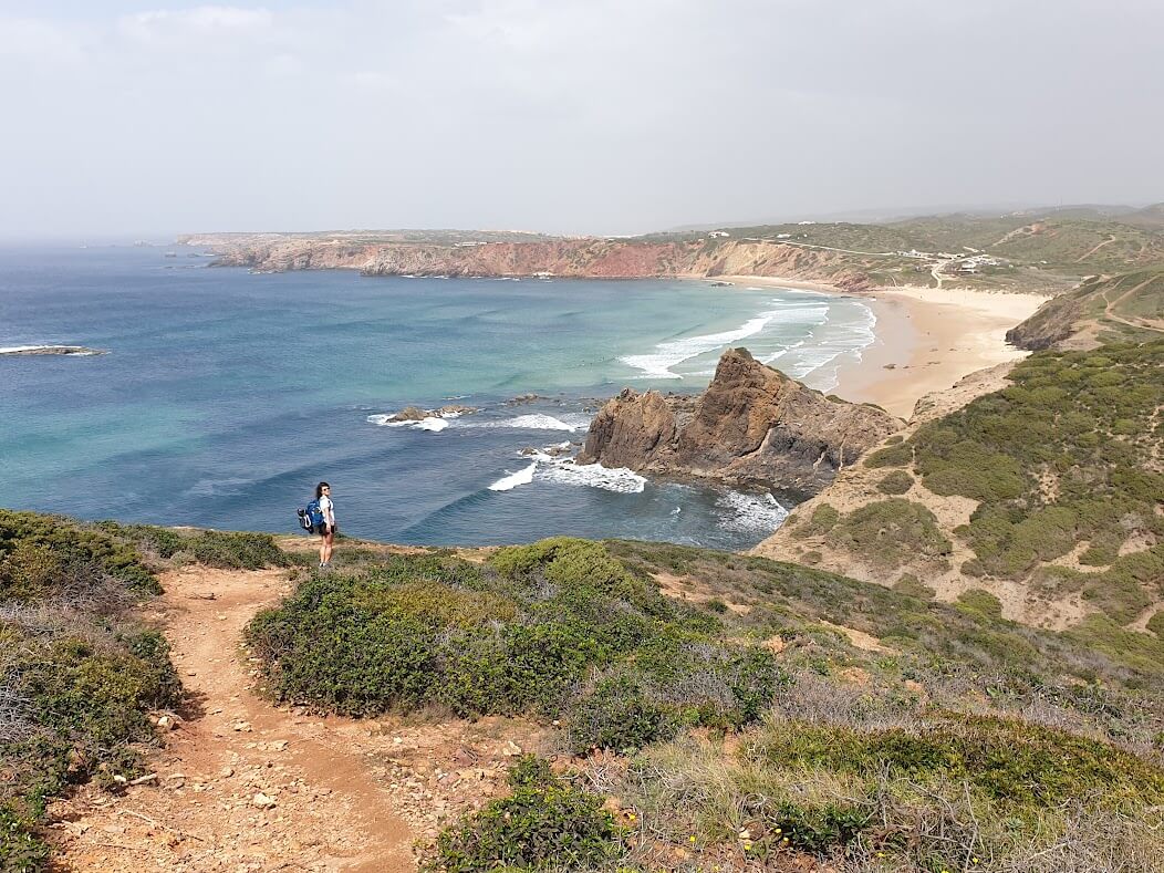 A woman with a blue rucksack stands on an orange-rock cliff. Below, a series of sandy beaches meet deep blue water, broken by white waves.