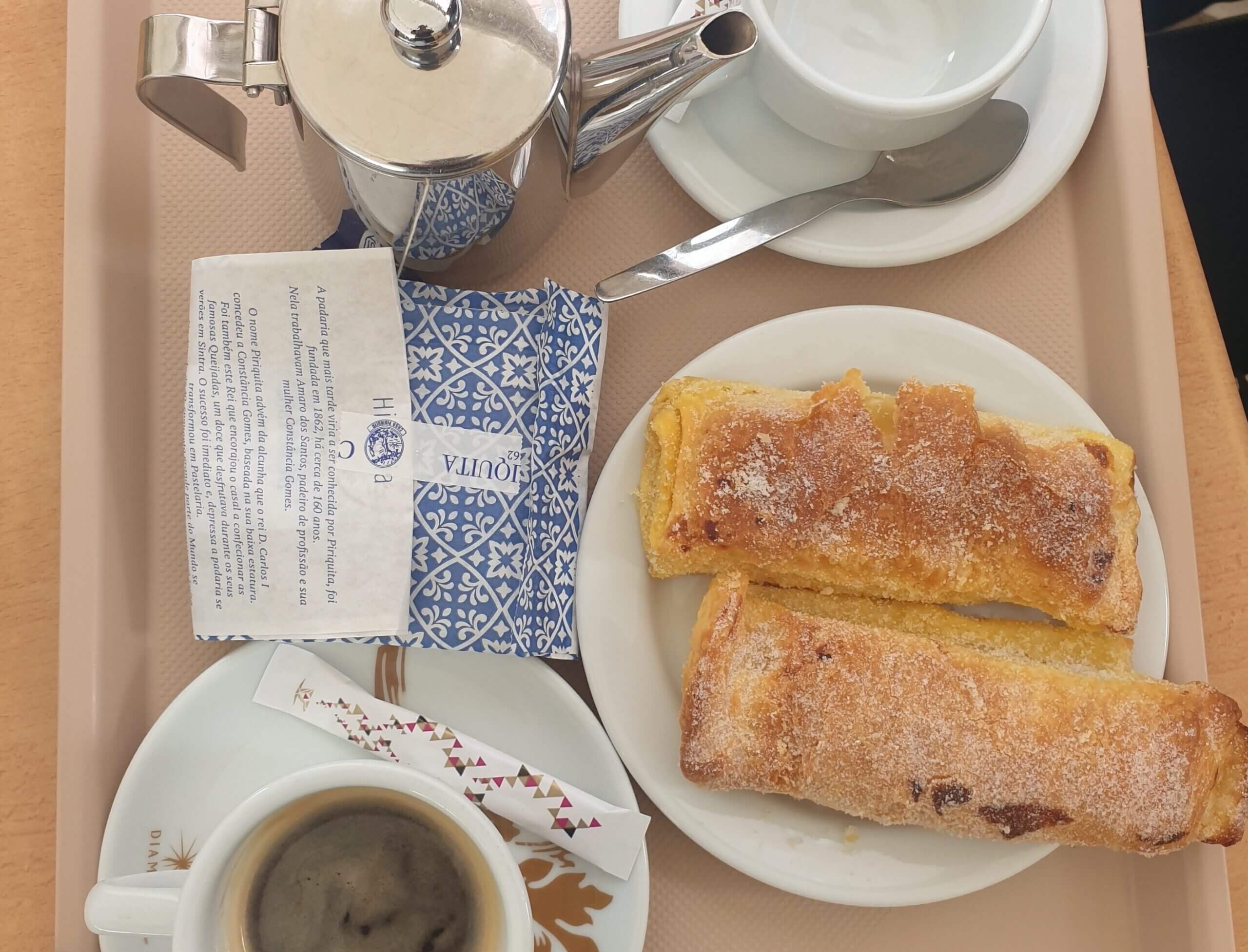 Bird's eye view shows two pillow-shaped pastries on a plate, a cup of black coffee, an empty cup and saucer, and a gleaming metal tea pot. All surround a blue and white sealed paper bag